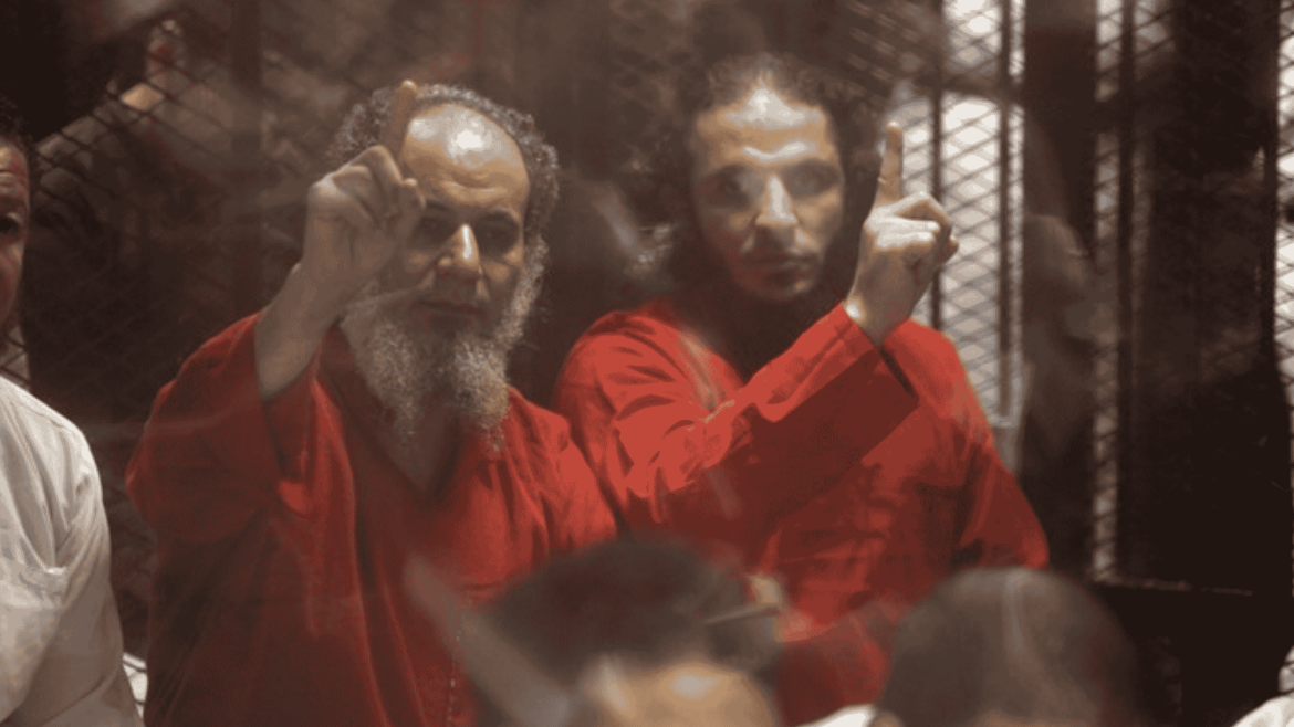 مشهد من داخل قاعة محكمة في مصر يُظهر عددًا من المتهمين يرتدون الزي الأحمر المخصص للمحكوم عليهم بالإعدام، يرفعون أيديهم بإشارات تعبر عن التحدي والإصرار، خلف قضبان قفص الاتهام.
