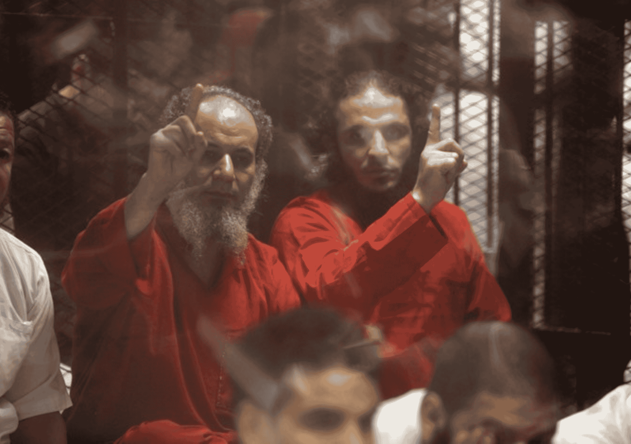 مشهد من داخل قاعة محكمة في مصر يُظهر عددًا من المتهمين يرتدون الزي الأحمر المخصص للمحكوم عليهم بالإعدام، يرفعون أيديهم بإشارات تعبر عن التحدي والإصرار، خلف قضبان قفص الاتهام.