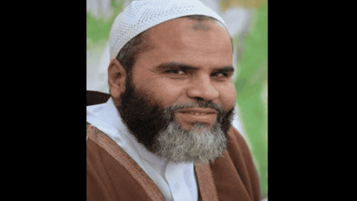 المعتقل فضل المولى، يرتدي جلبابًا بنيًا وغطاء رأس أبيض، يبتسم ابتسامة خفيفة أثناء التقاط الصورة في مكان مفتوح يبدو أن به خلفية خضراء غير واضحة.