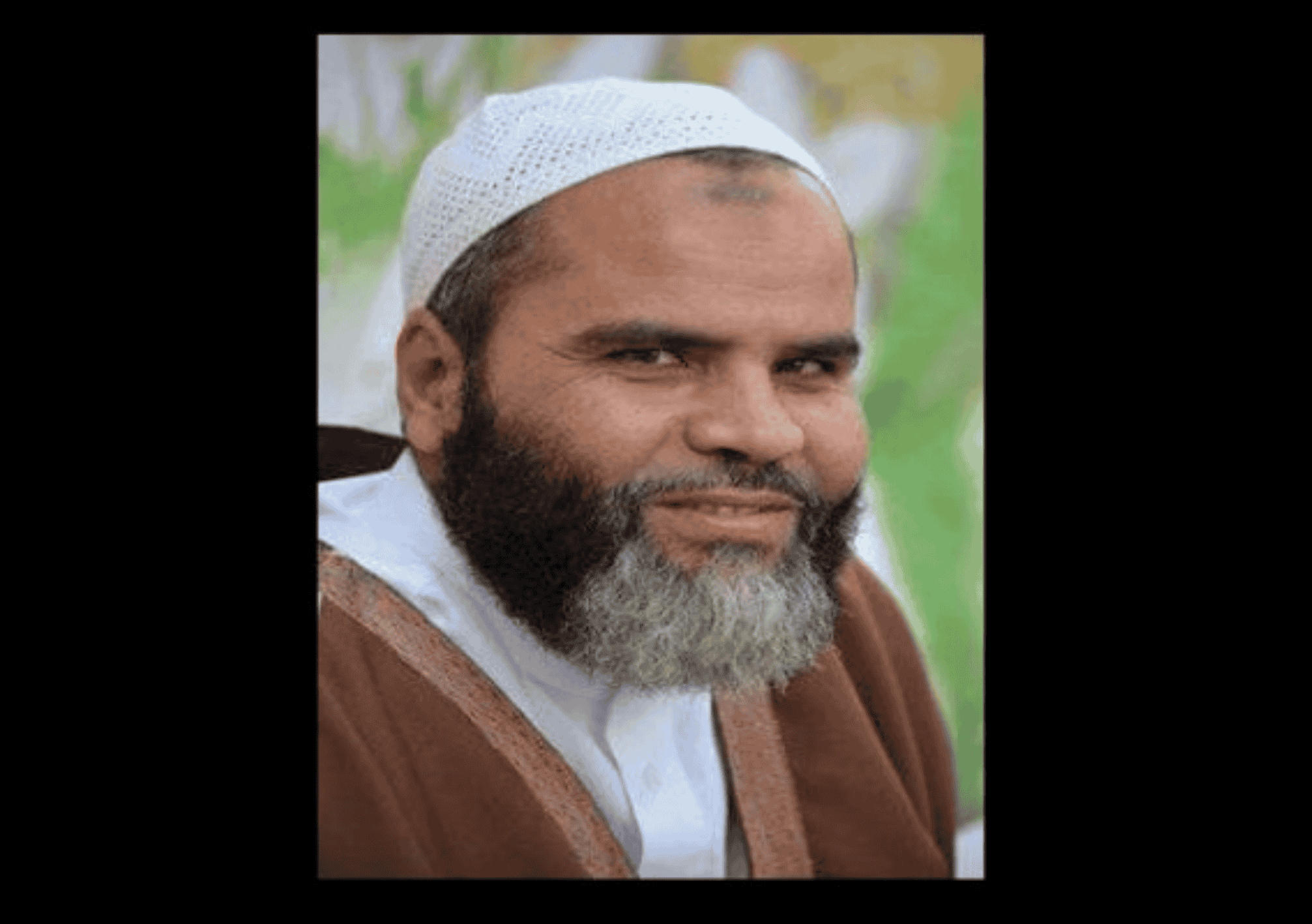 المعتقل فضل المولى، يرتدي جلبابًا بنيًا وغطاء رأس أبيض، يبتسم ابتسامة خفيفة أثناء التقاط الصورة في مكان مفتوح يبدو أن به خلفية خضراء غير واضحة.