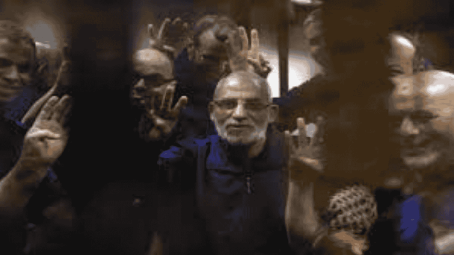 متهمون سياسيون يرفعون شعار رابعة داخل قفص زجاجي في محكمة مصرية.