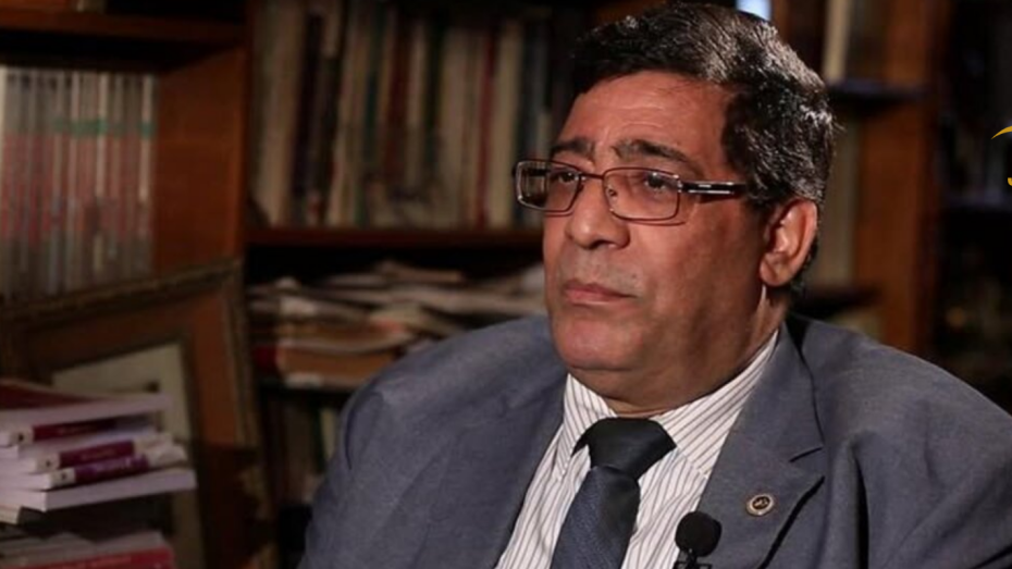 رجل يرتدي بدلة رسمية رمادية ونظارة يتحدث داخل مكتبة مليئة بالكتب، أثناء مقابلة تلفزيونية.