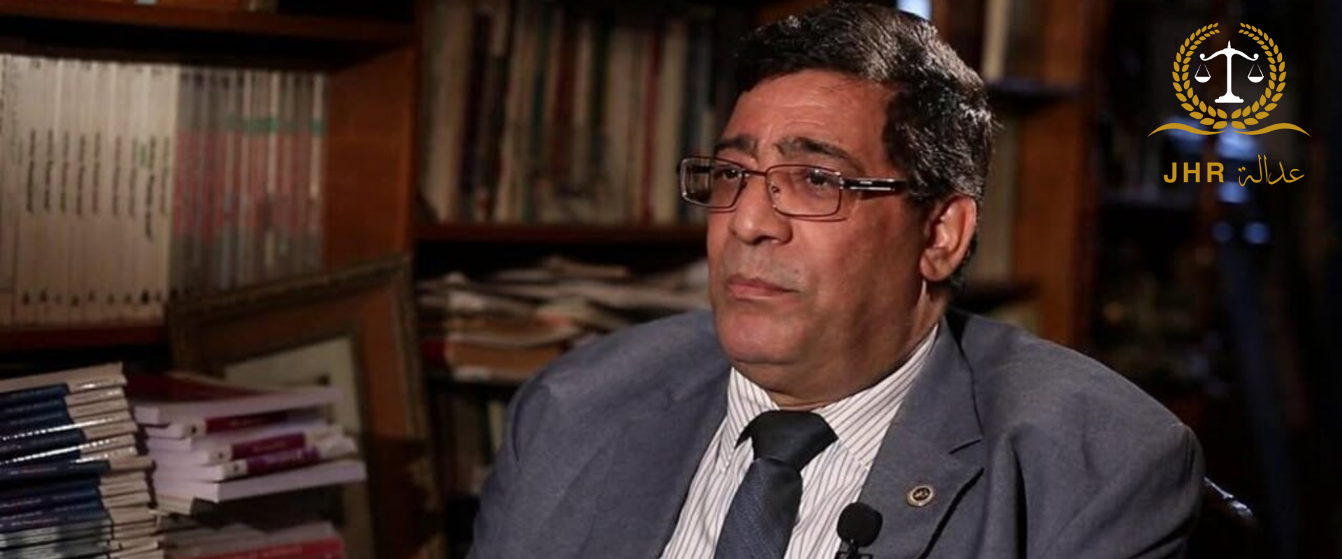 رجل يرتدي بدلة رسمية رمادية ونظارة يتحدث داخل مكتبة مليئة بالكتب، أثناء مقابلة تلفزيونية.