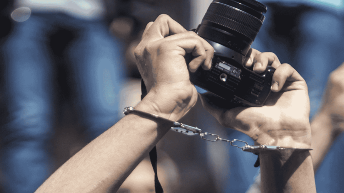 يد صحفي مقيدة بالقيود وهو يرفع كاميرا تصوير أثناء عمله، في رمز لقمع حرية الصحافة.