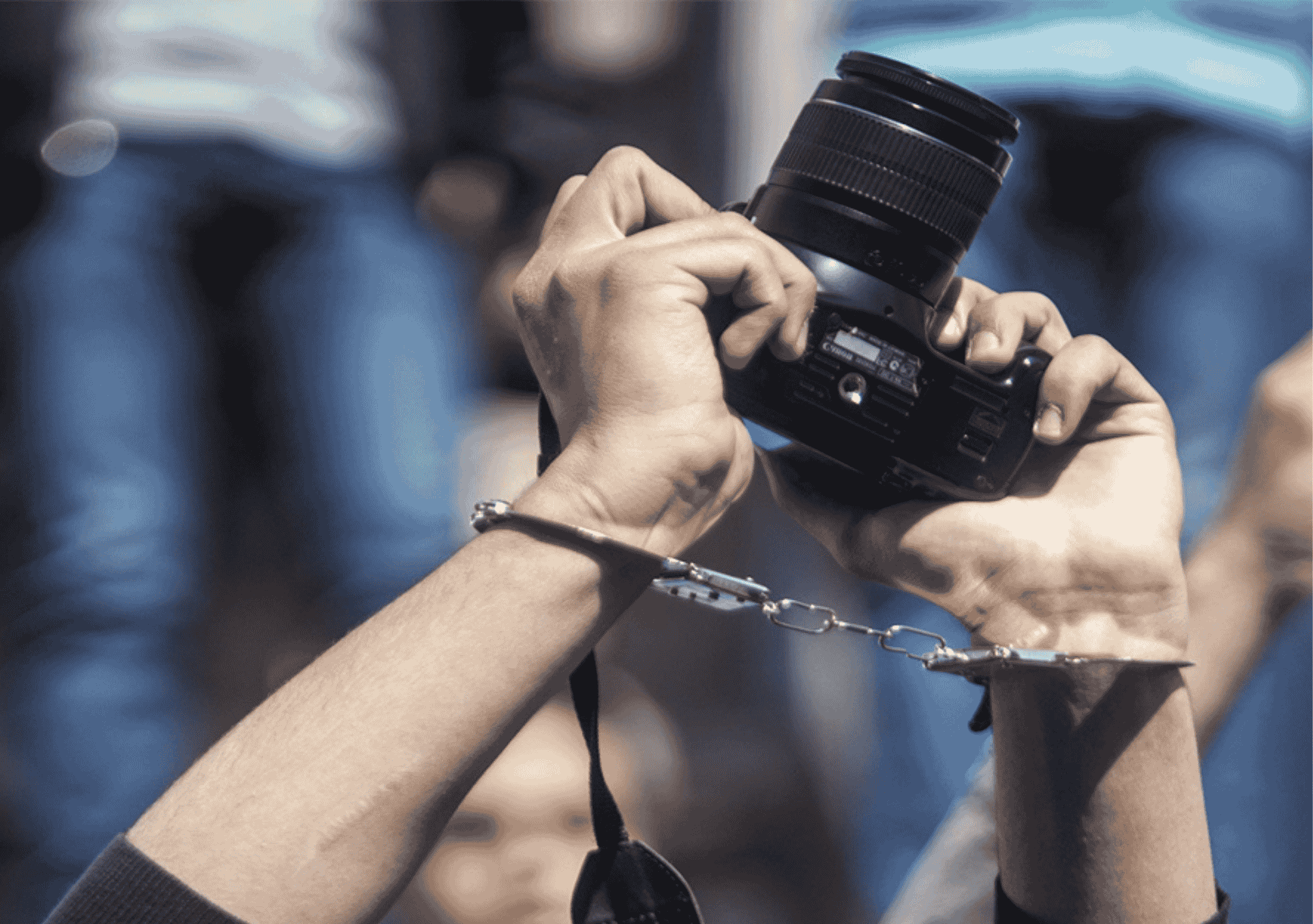 يد صحفي مقيدة بالقيود وهو يرفع كاميرا تصوير أثناء عمله، في رمز لقمع حرية الصحافة.