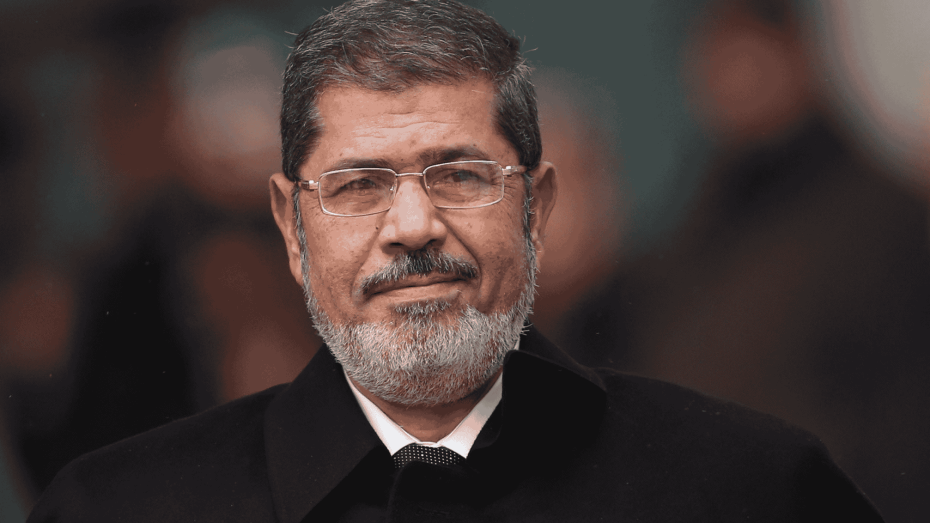 الرئيس الشهيد محمد مرسي يرتدي معطفًا أسود ونظارة طبية، يقف في مكان عام ويبدو بنظرة هادئة.