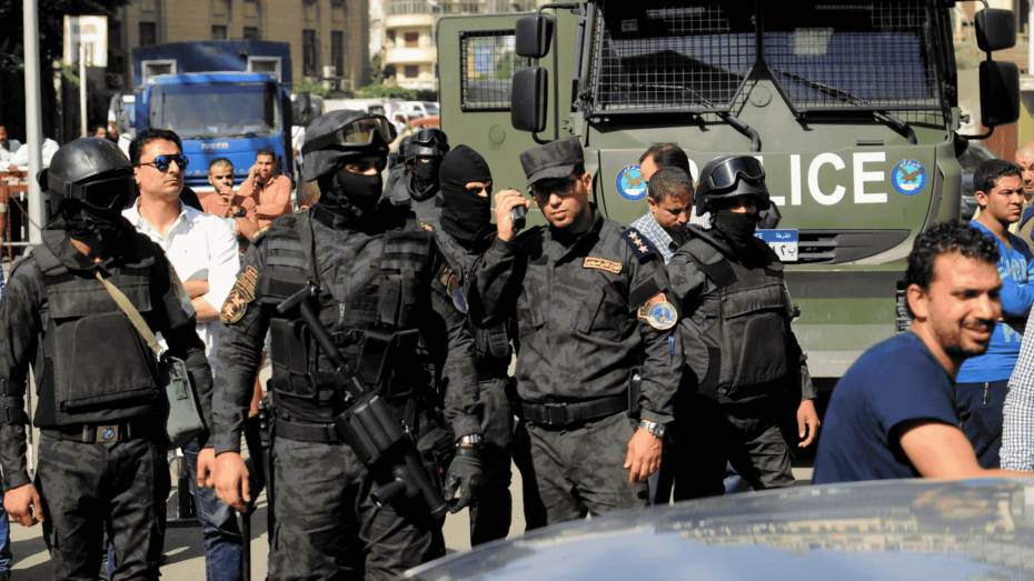 مجموعة من أفراد قوات الأمن المصرية يرتدون الزي الأسود المخصص للعمليات الخاصة، ويقفون أمام مدرعة شرطة في أحد شوارع القاهرة، وسط تواجد مدنيين ومركبات أمنية.