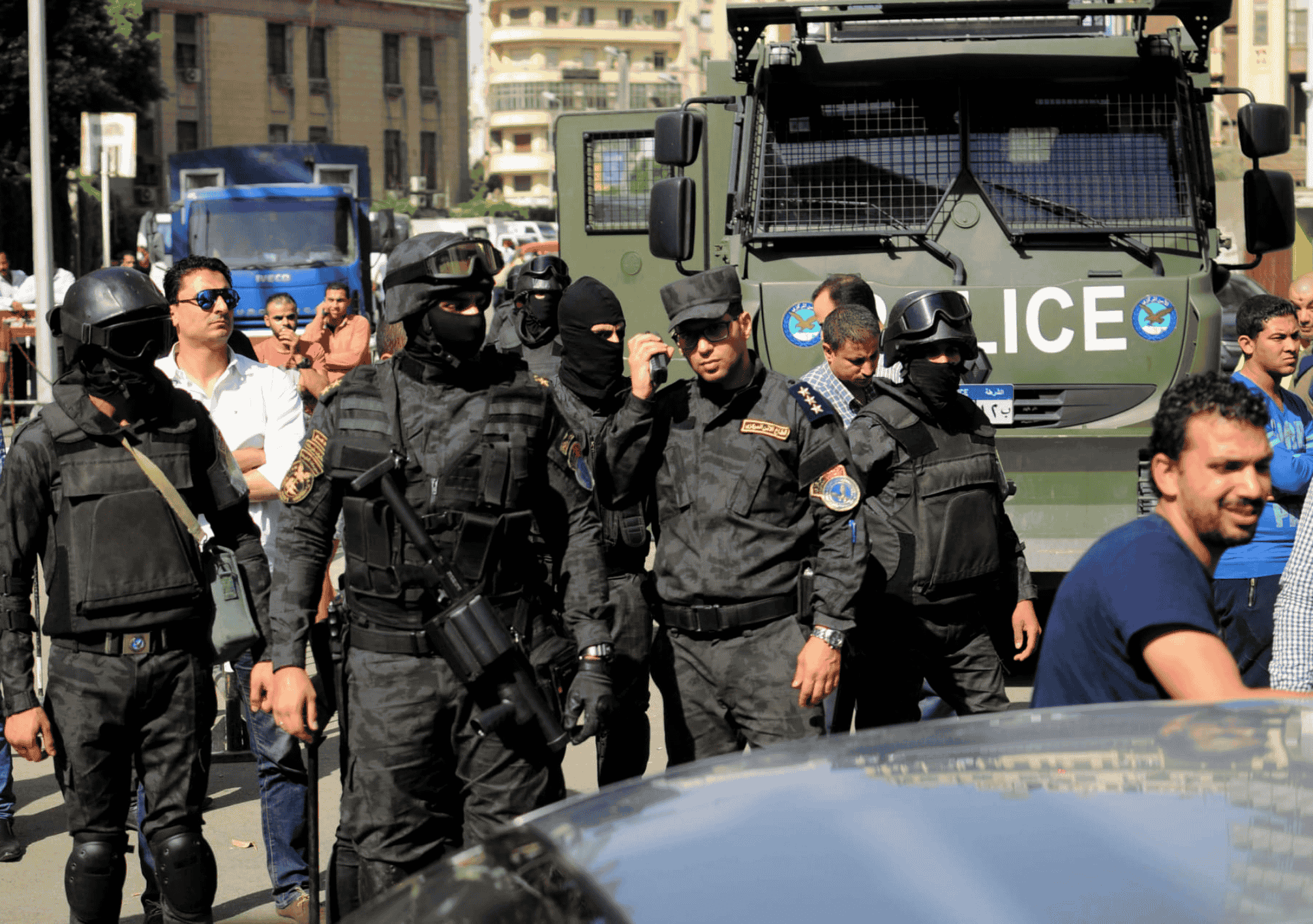 مجموعة من أفراد قوات الأمن المصرية يرتدون الزي الأسود المخصص للعمليات الخاصة، ويقفون أمام مدرعة شرطة في أحد شوارع القاهرة، وسط تواجد مدنيين ومركبات أمنية.