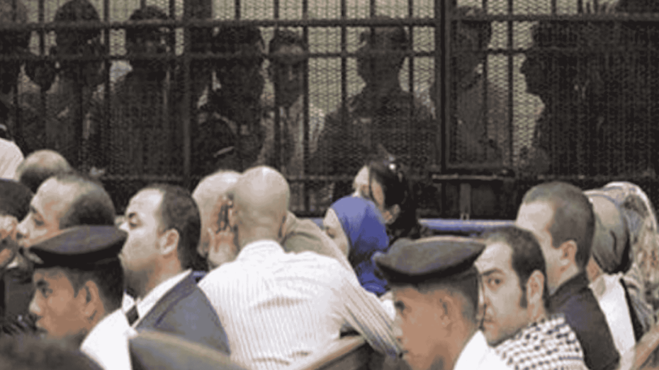 صورة من داخل قاعة محكمة مصرية تُظهر متهمين يجلسون داخل قفص الاتهام الحديدي، وخلفهم حراسة مشددة من قوات الأمن، في مشهد يعكس واقع المحاكمات الجماعية في مصر.