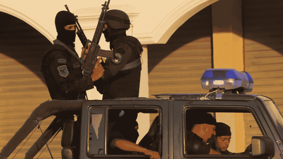 صورة تظهر مجموعة من عناصر الأمن المسلحين يرتدون الزي الأسود والأقنعة الواقية، يقفون على متن عربة شرطة مجهزة بضوء الطوارئ الأزرق، في مشهد يعكس الانتشار الأمني المكثف في الشوارع المصرية.