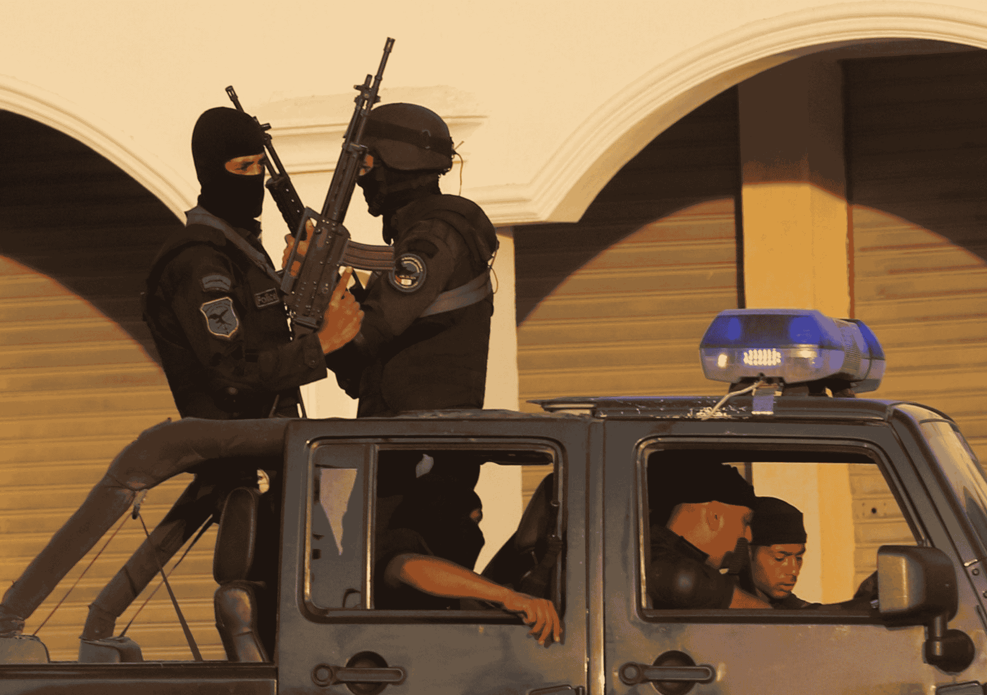 صورة تظهر مجموعة من عناصر الأمن المسلحين يرتدون الزي الأسود والأقنعة الواقية، يقفون على متن عربة شرطة مجهزة بضوء الطوارئ الأزرق، في مشهد يعكس الانتشار الأمني المكثف في الشوارع المصرية.