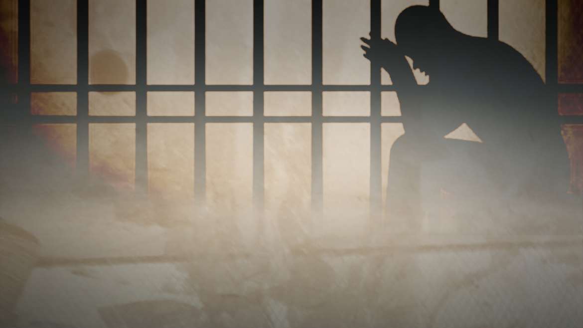 تُظهر الصورة ظلاً لرجل يجلس خلف قضبان حديدية، واضعاً رأسه بين يديه في وضعٍ يدل على الحزن واليأس، داخل زنزانة يغمرها ضوء خافت من الخارج. يرمز المشهد إلى حالات الاعتقال غير القانوني والإخفاء القسري وما يرافقها من معاناة إنسانية وانتهاك للكرامة.